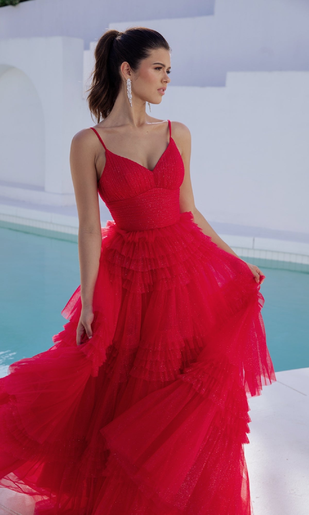 A woman stands by a pool in the Eureka 9888 Lace-Up Long Glitter-Tulle Prom Dress, holding her bright red gown with one hand; white walls and arches form the background.
